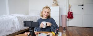 A young woman smiling at a camera while holding a mug, seated at a makeup table with various beauty products, in a cozy bedroom setup, recording a video.