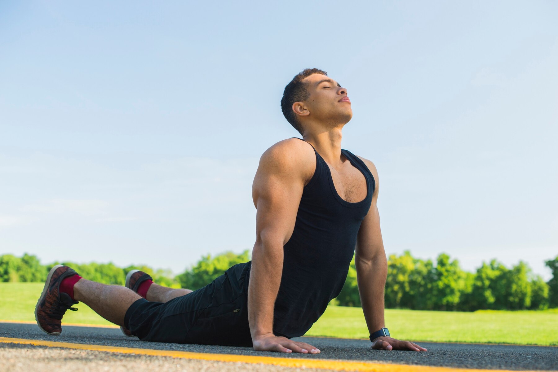A young man performing an upward-facing stretch on a sunny road with a grassy field and clear sky in the background.