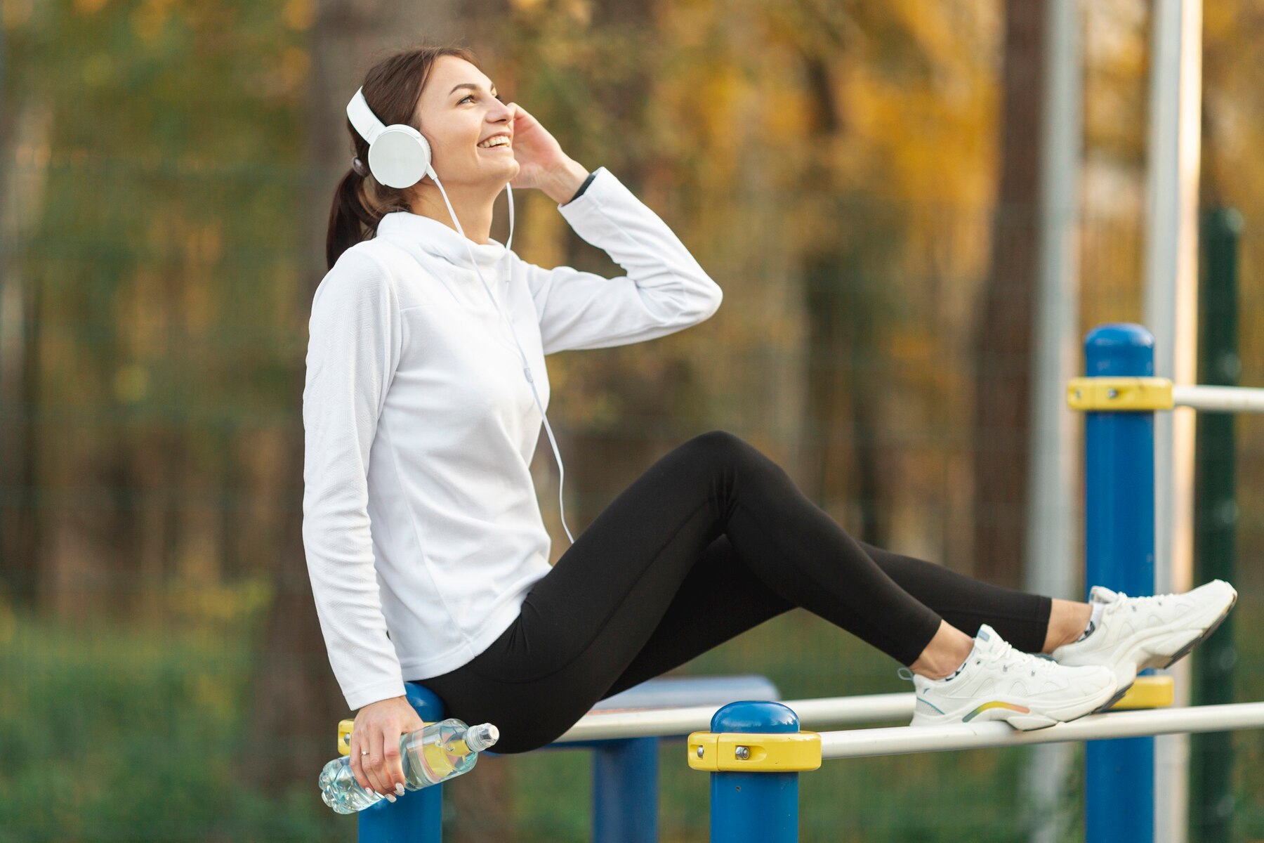 A joyful woman in sportswear and headphones sits on a horizontal bar at an outdoor gym, holding a water bottle, with a blurred autumnal background.