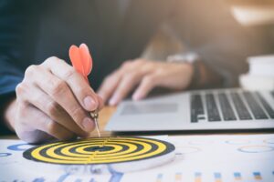 A hand holding a dart poised to hit a target on a desk with a laptop and documents, symbolizing focus and goal achievement.