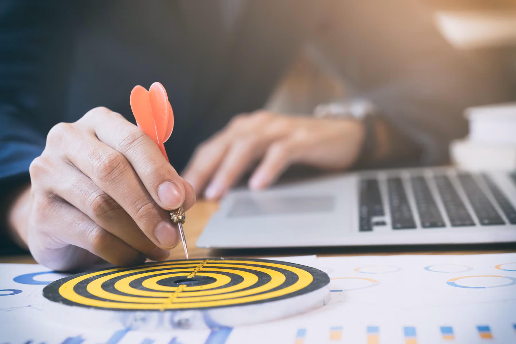A hand holding a dart poised to hit a target on a desk with a laptop and documents, symbolizing focus and goal achievement.