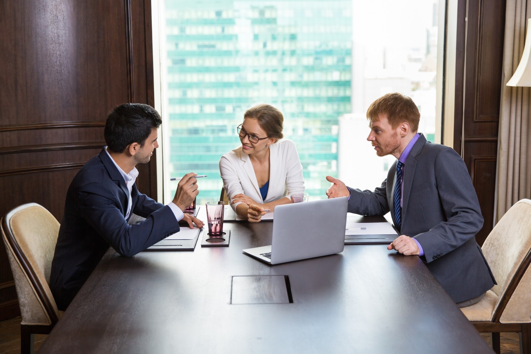 Three professionals engaged in a discussion around a conference table in an office with a cityscape visible through the window.