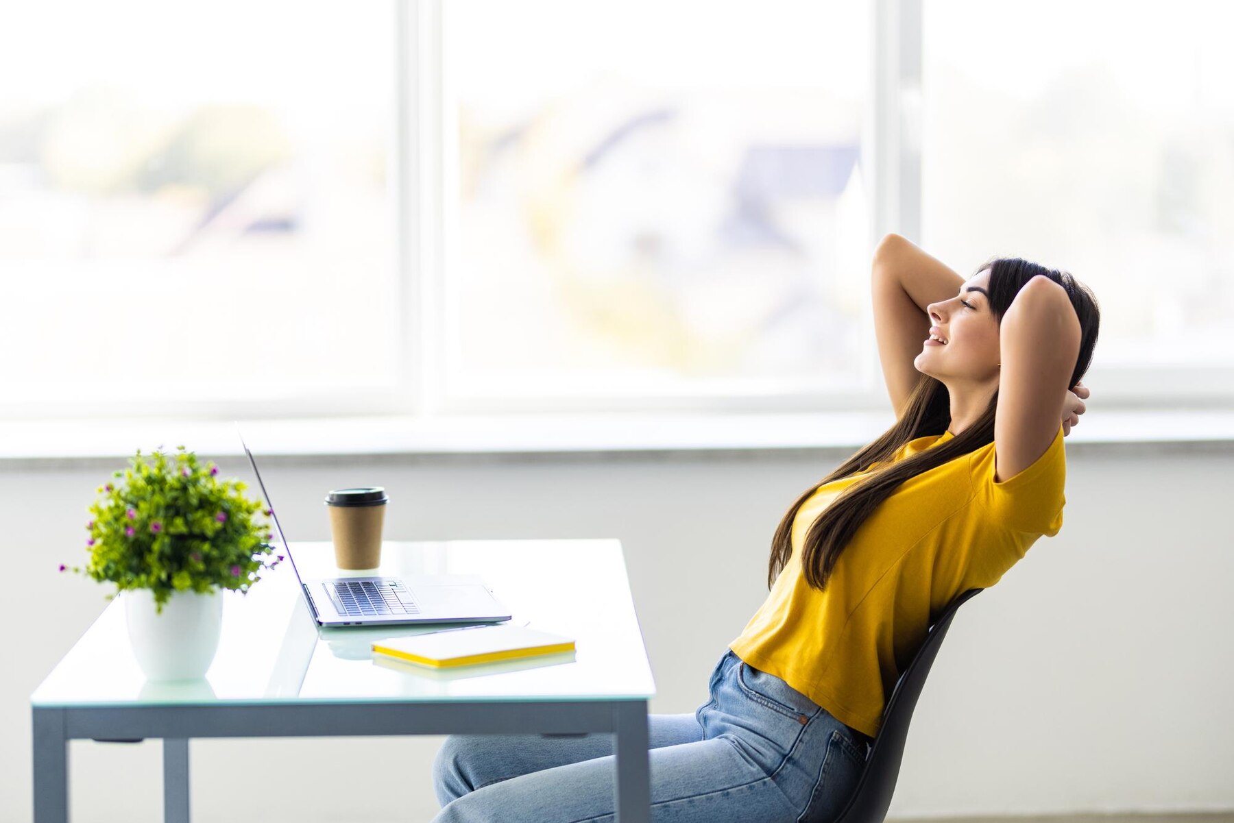 A woman in a yellow top and jeans sitting relaxed in an office chair, hands clasped behind her head, next to a desk with a laptop, a notebook, and a coffee cup, in a brightly lit room.