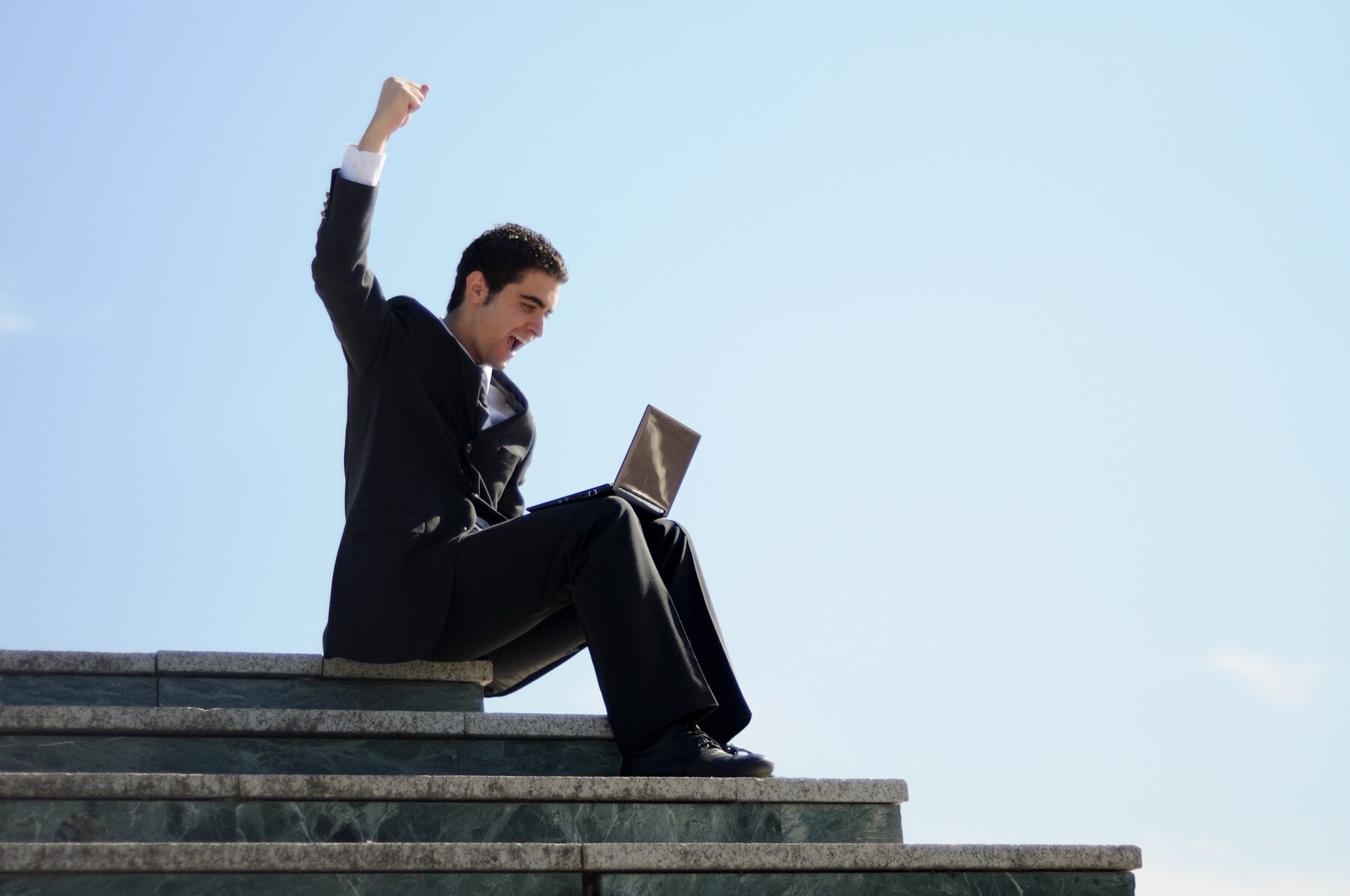 Businessman in a suit sitting on steps outside, triumphantly raising his arm while looking at a laptop.
