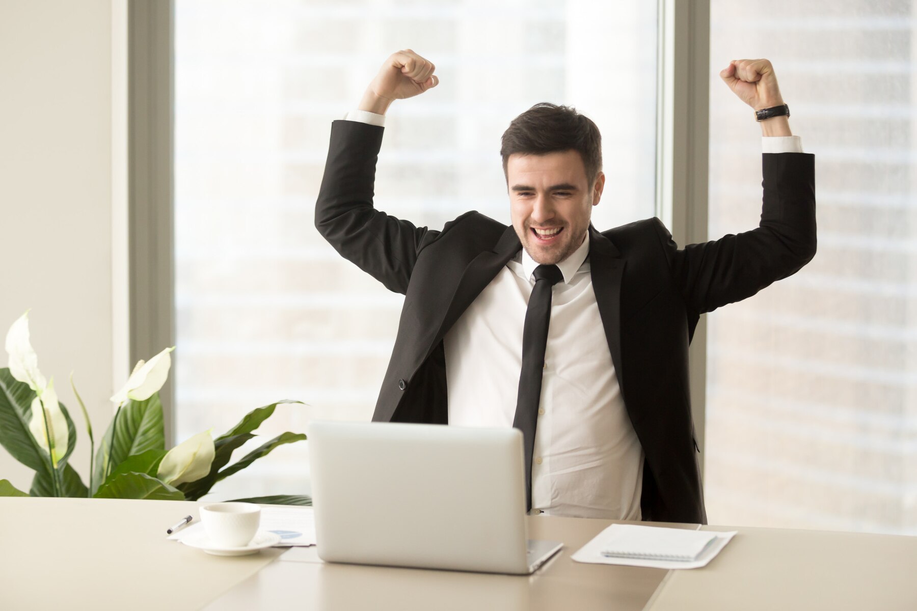 A joyful businessman celebrating success at his desk with a laptop, fists raised in victory.