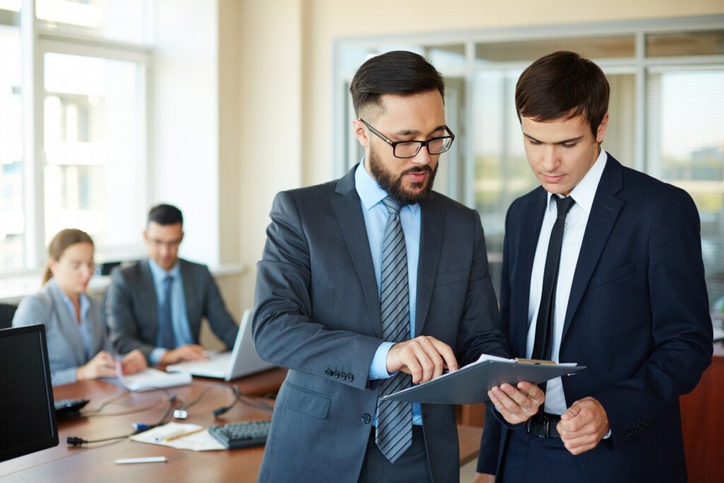 Two businessmen discussing documents on a tablet in a modern office setting, with colleagues working in the background.