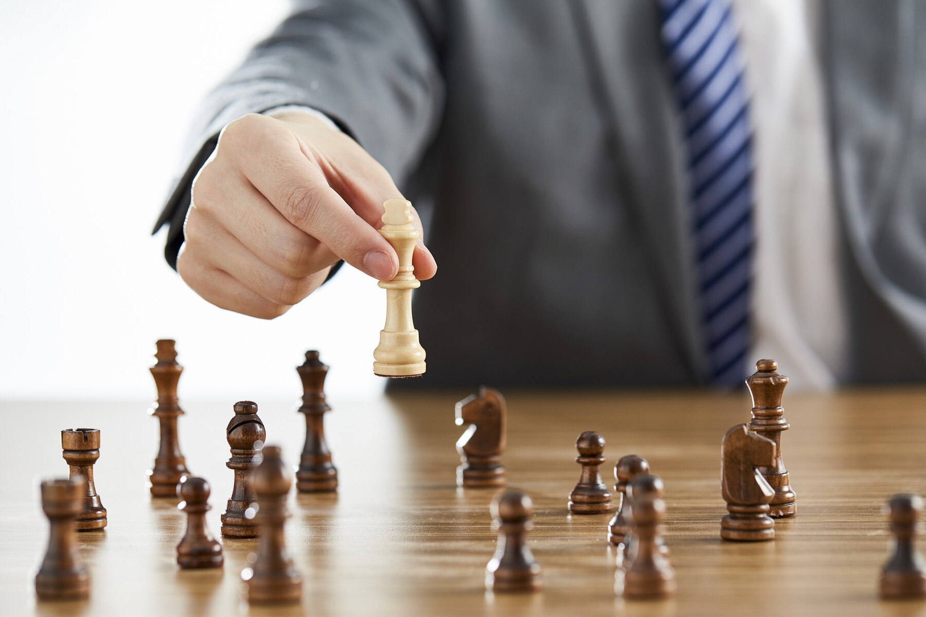 A person in a suit moves a white king chess piece in a game against brown pieces on a wooden table.