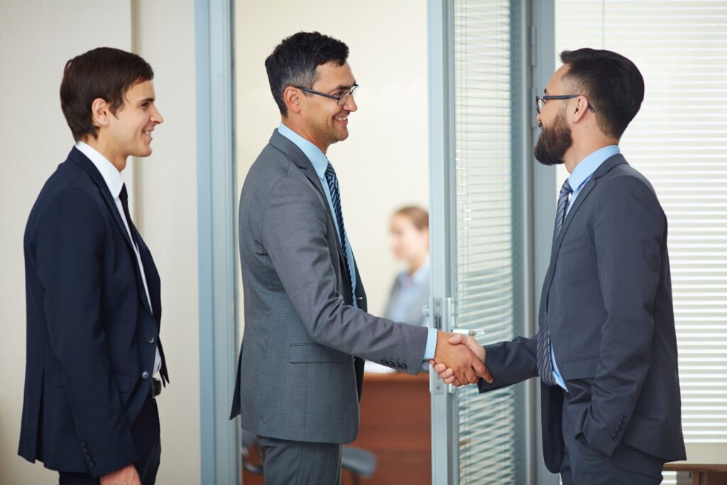 Two business professionals shake hands in an office setting, while a third person observes nearby. Modern furnishings are visible.