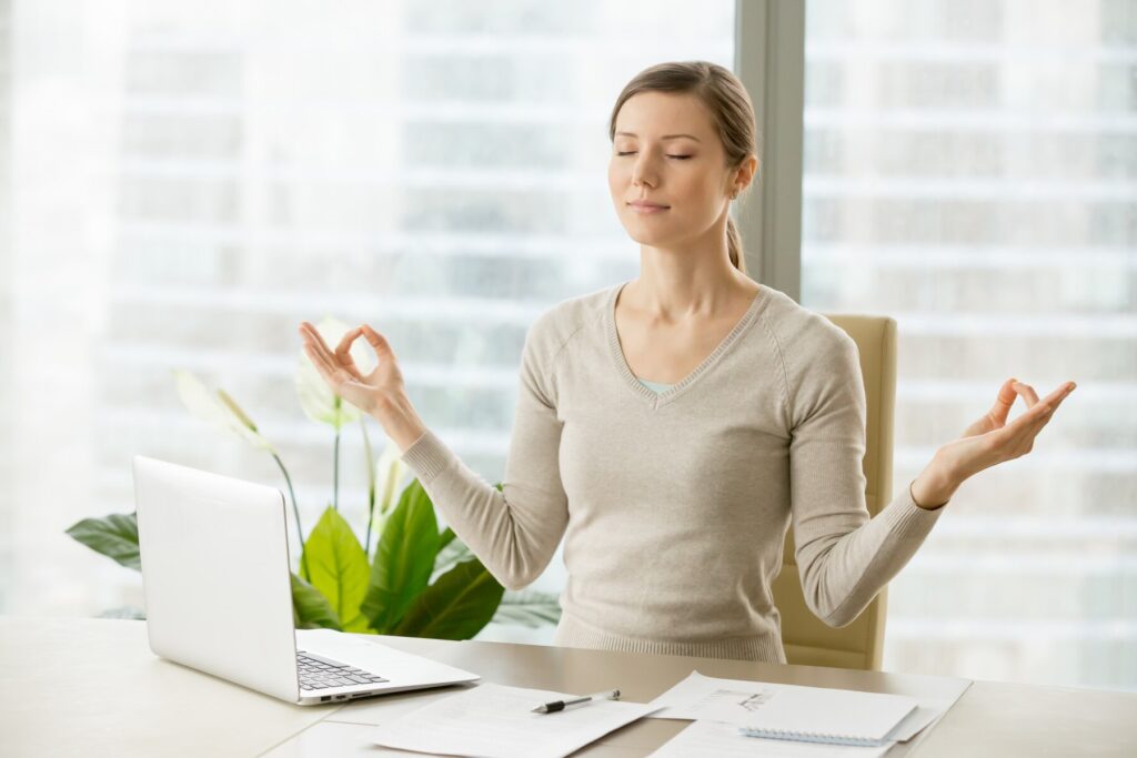 A woman meditating at her office desk with a laptop and documents, practising stress-relief techniques in a bright workspace.