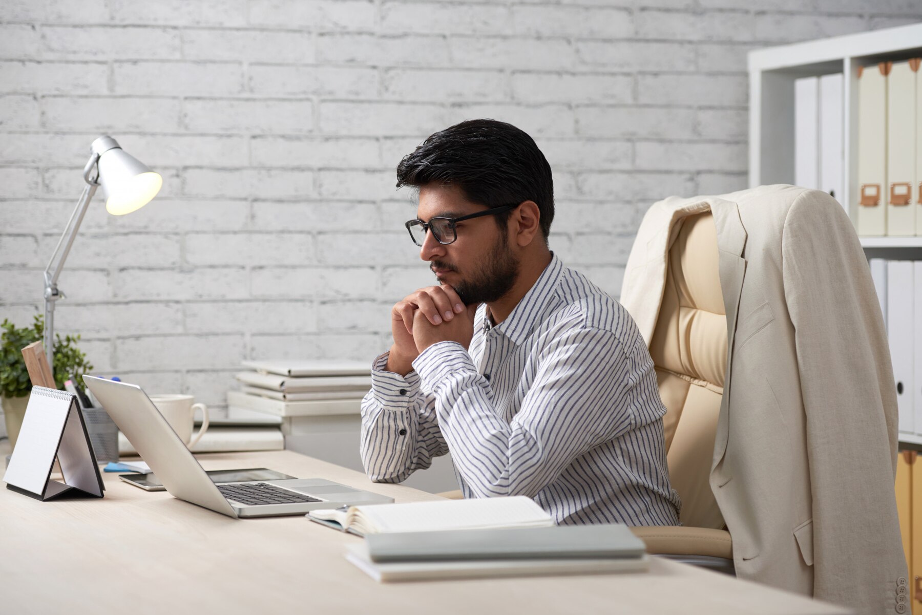 A person sits at a desk with a laptop, a stack of books, and a desk lamp, deep in thought in a modern office setting.