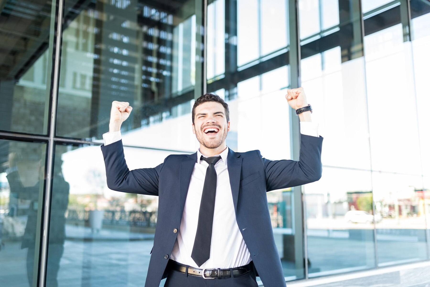 A jubilant young man in a suit with raised fists celebrating success outside a modern glass building.