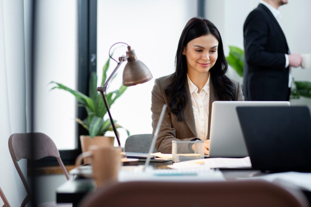 A businesswoman using a laptop at a modern office desk, smiling while looking at the screen, with a colleague in the background.