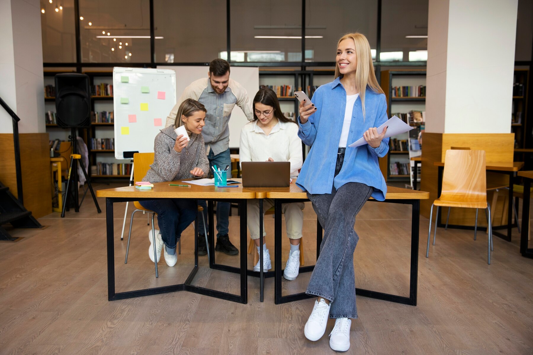 Two women and two men collaborate around a wooden table in a modern office space, with one woman standing and using her smartphone, while the others discuss notes and a laptop.