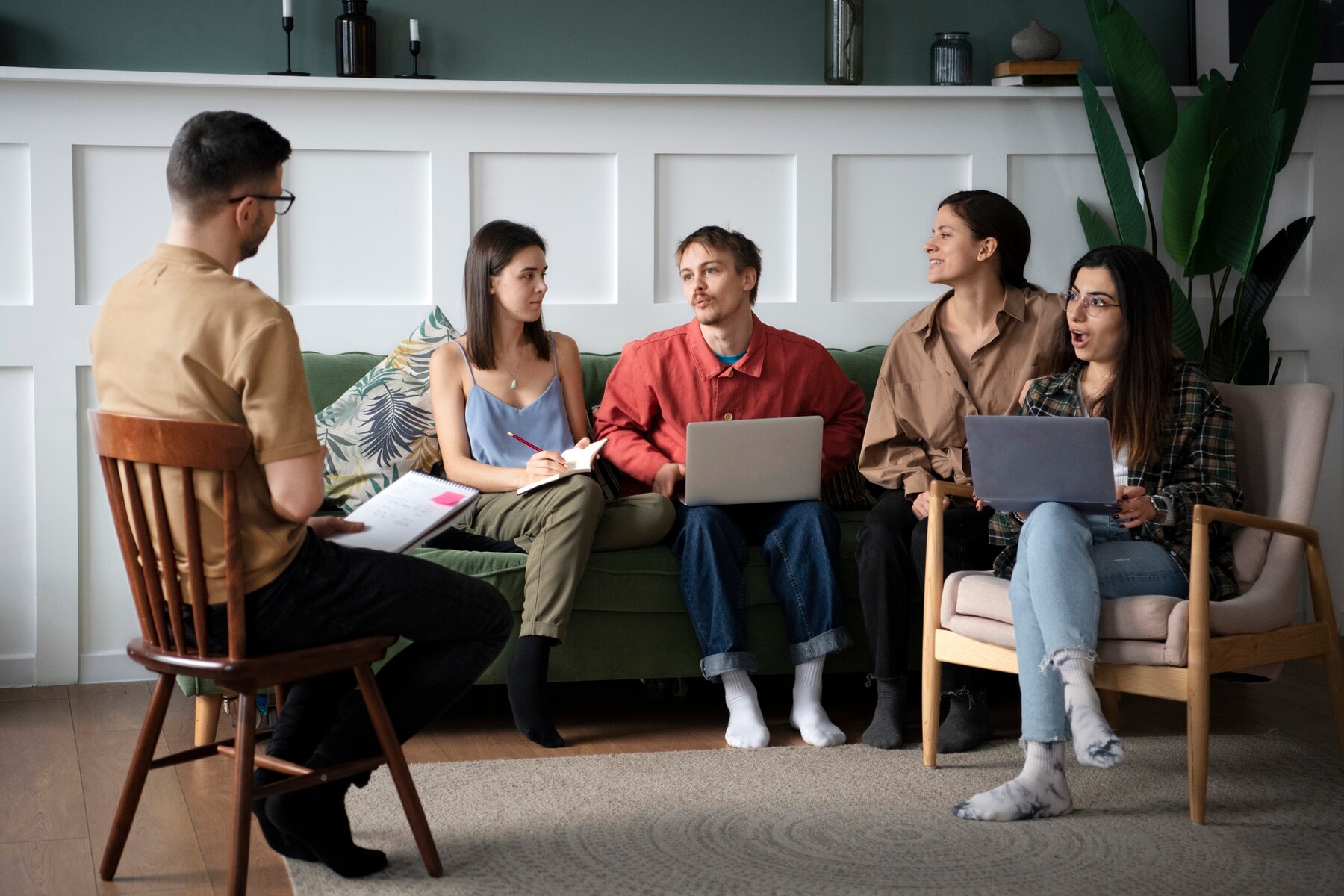 A group of young adults engaging in a discussion in a modern, stylish living room, with one man leading while seated on a chair facing four others on a couch and chairs, showcasing a focused, collaborative atmosphere.