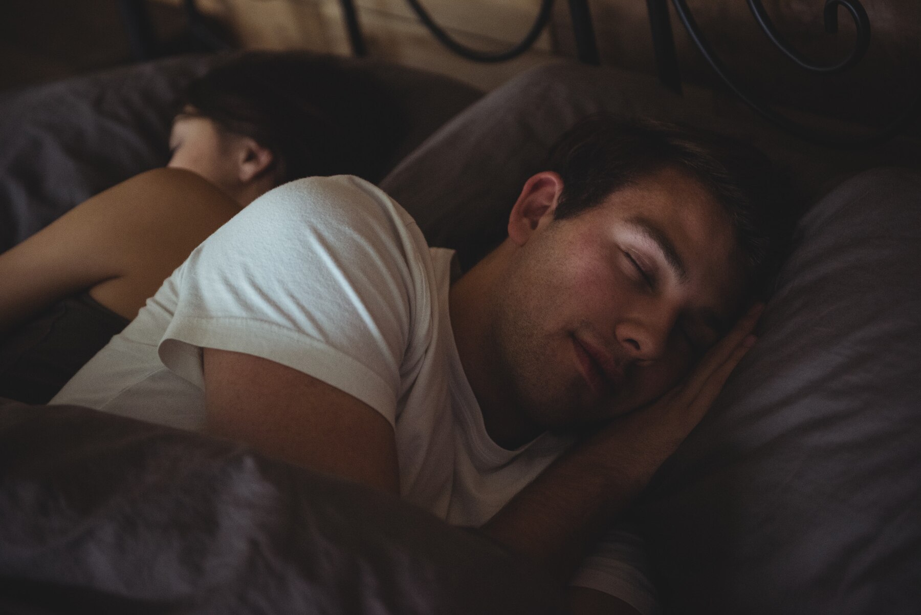 A young man and a woman sleeping peacefully in a dimly lit bedroom, with the man resting his head on his hands and the woman turned away from the camera.