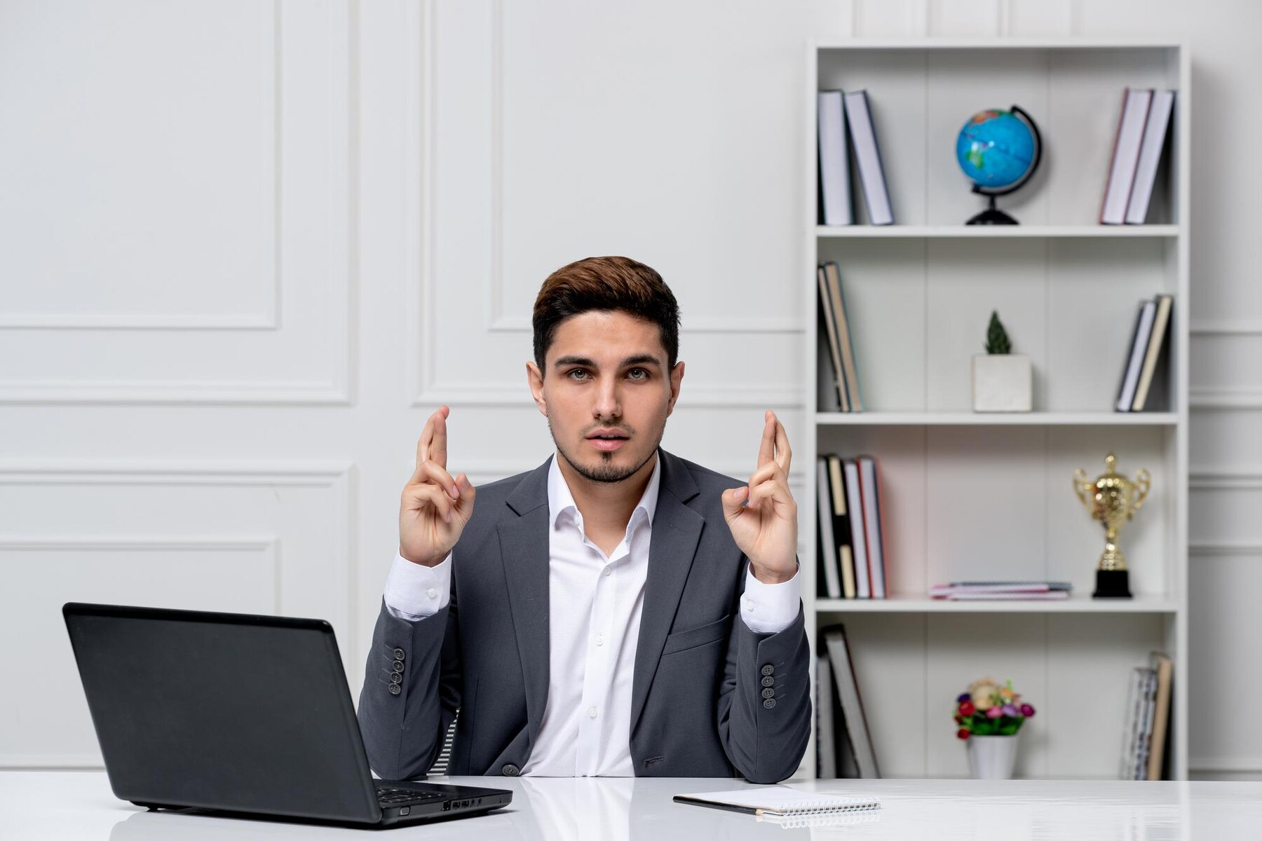 A young businessman in a grey suit sitting at his desk with a laptop, crossing his fingers for luck, in a modern office with a bookshelf in the background.