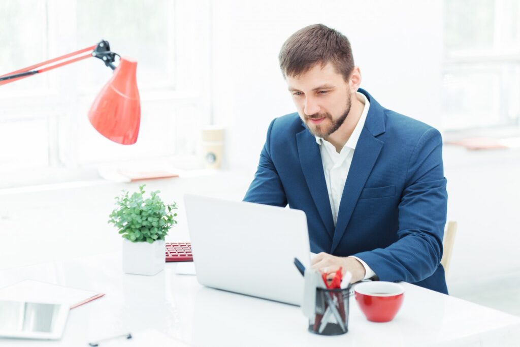 A businessman in a blue suit working intently on a laptop at a bright office desk, accompanied by a red desk lamp, a small green plant, and a red mug.