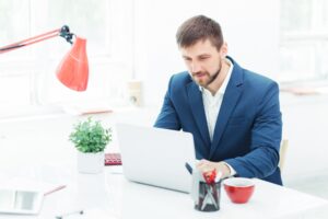 A businessman in a blue suit working intently on a laptop at a bright office desk, accompanied by a red desk lamp, a small green plant, and a red mug.