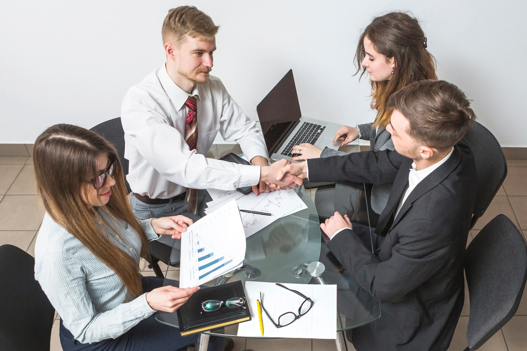 Four professionals engaged in a business meeting, shaking hands over a glass table with laptops and documents.