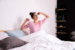 A person in a pink shirt stretches in bed surrounded by white blankets and pillows, with a modern shelf in the background.