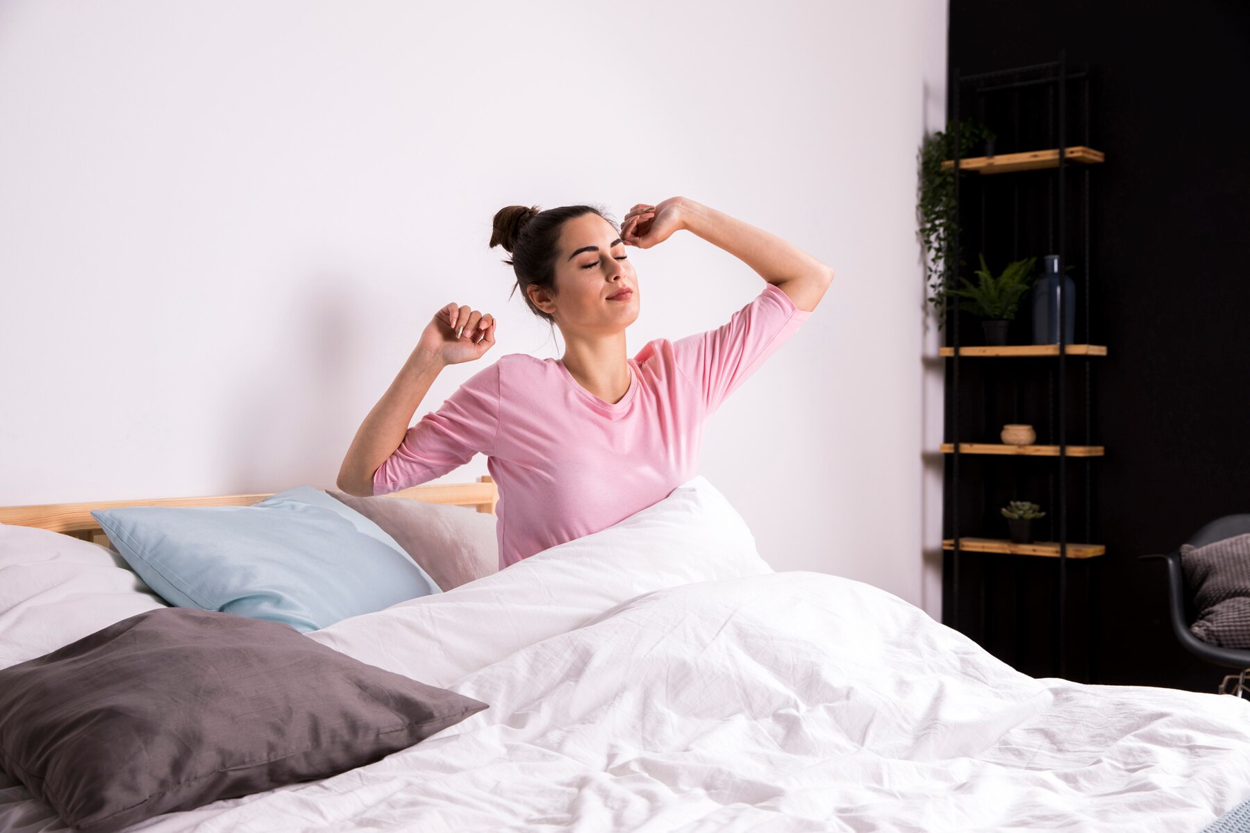 A person in a pink shirt stretches in bed surrounded by white blankets and pillows, with a modern shelf in the background.