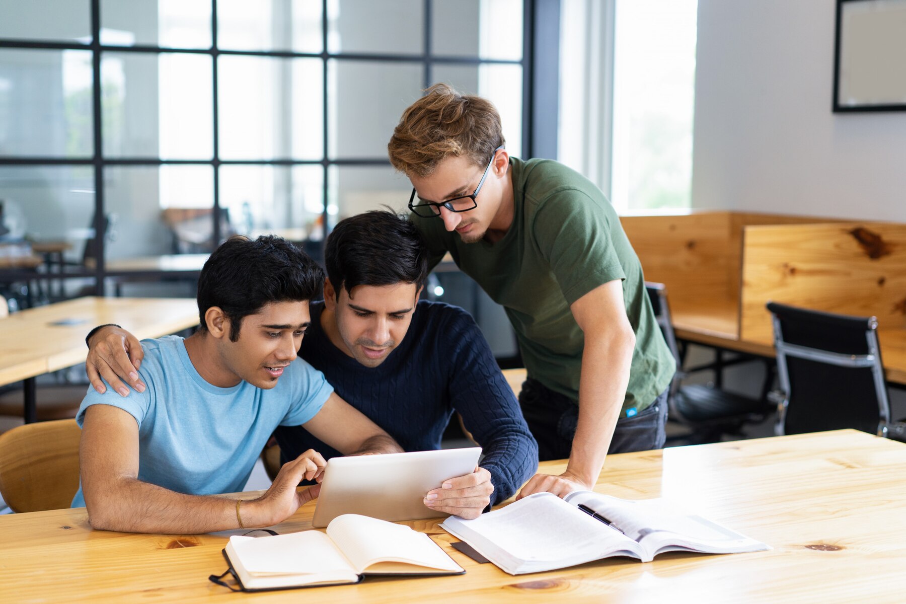 Three men collaboratively studying with digital tablet and books at a wooden table in a modern office with large windows.