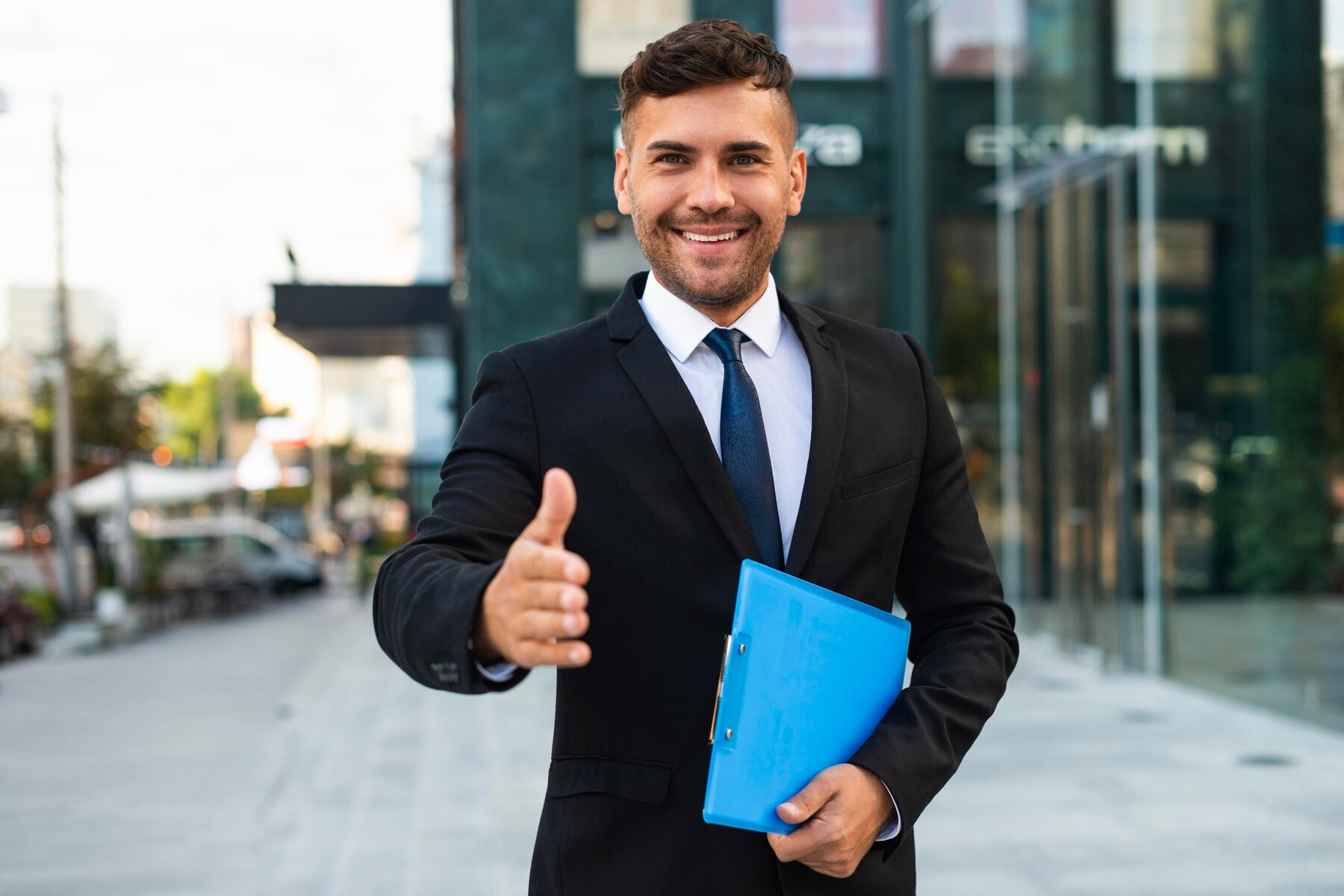 A confident young businessman in a suit with a blue tie, holding a blue folder and giving a thumbs up, standing on a city street.