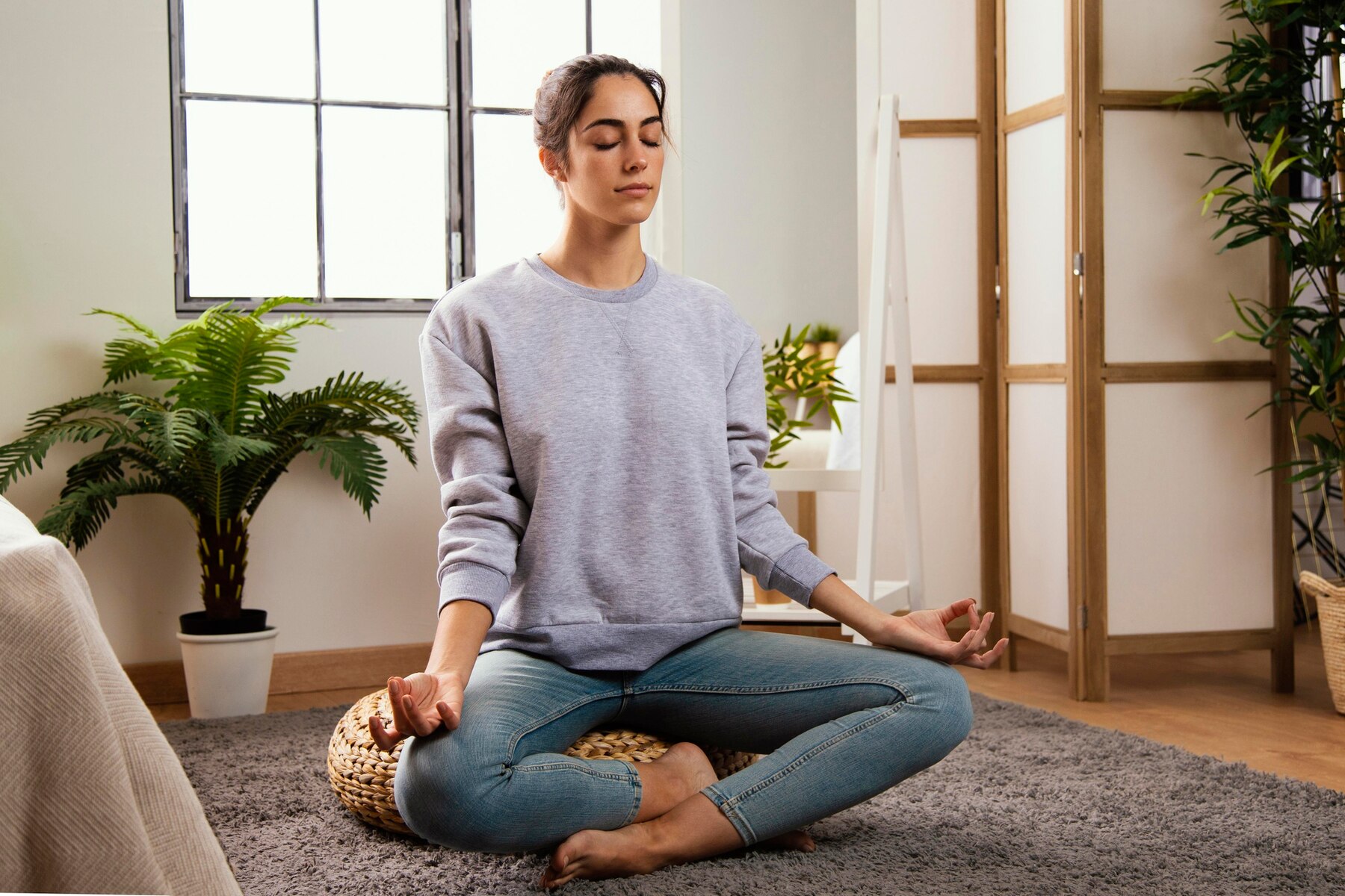 A woman meditating in a peaceful indoor setting, sitting cross-legged on a woven mat with closed eyes and upturned palms, surrounded by plants and natural light.