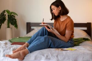 A young woman in casual attire sits cross-legged on a bed, focused on writing in a notebook, with a potted plant in the background.