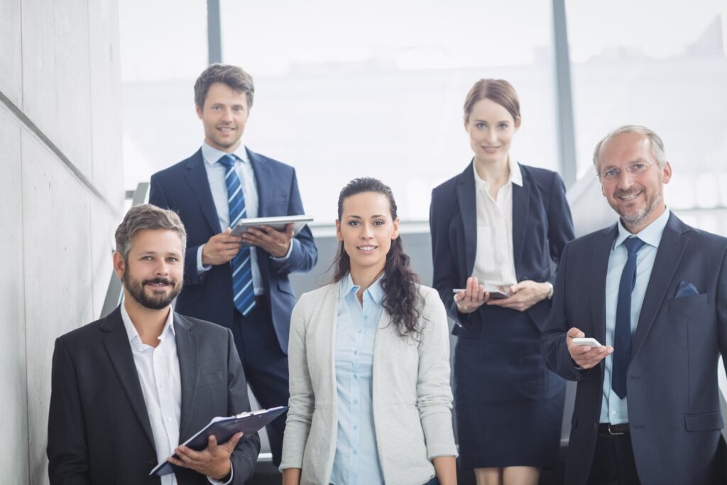 A group of five professionals in business attire, standing and engaging in conversation in a modern office environment.