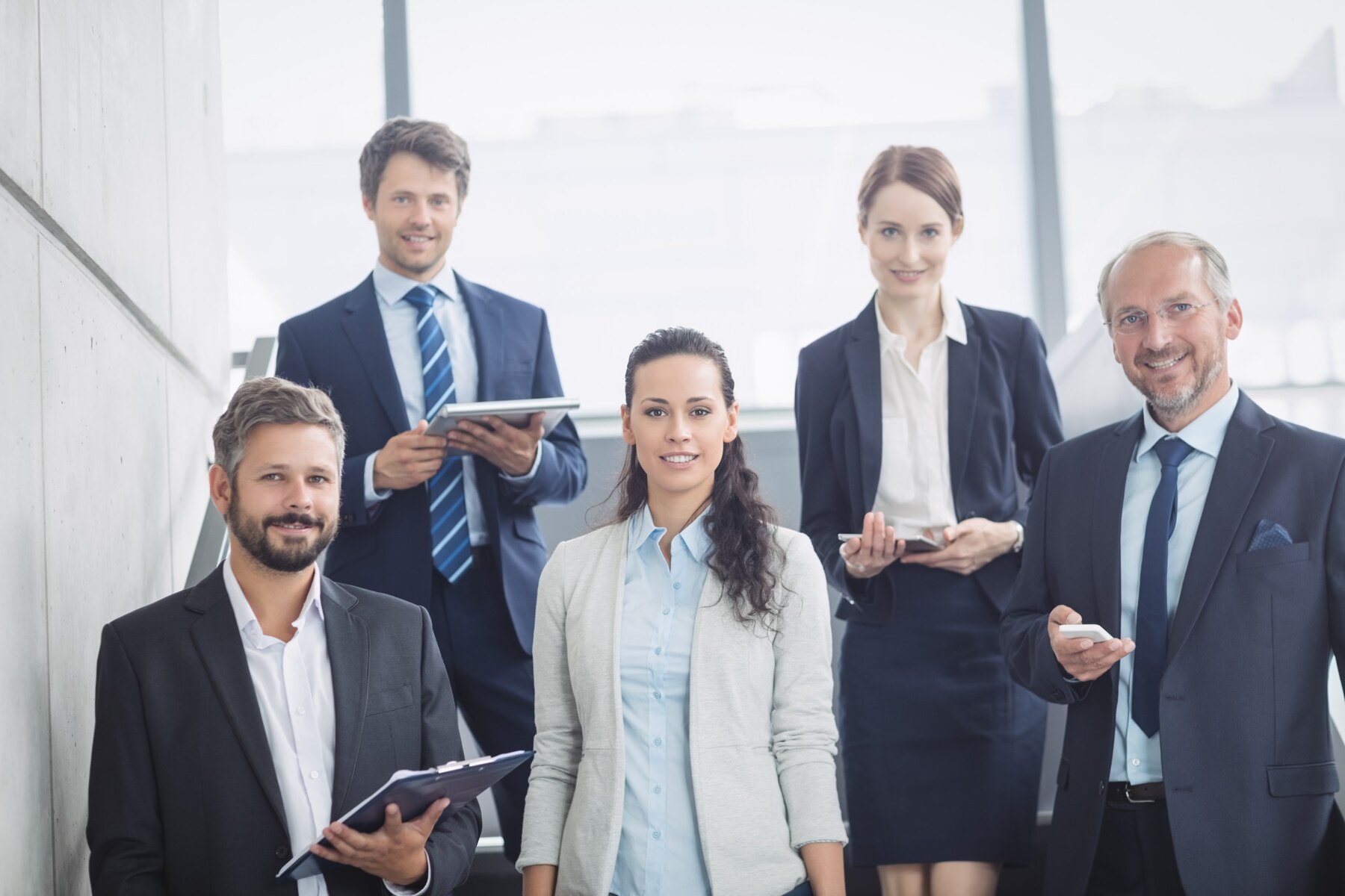 A group of five professionals in business attire, standing and engaging in conversation in a modern office environment.