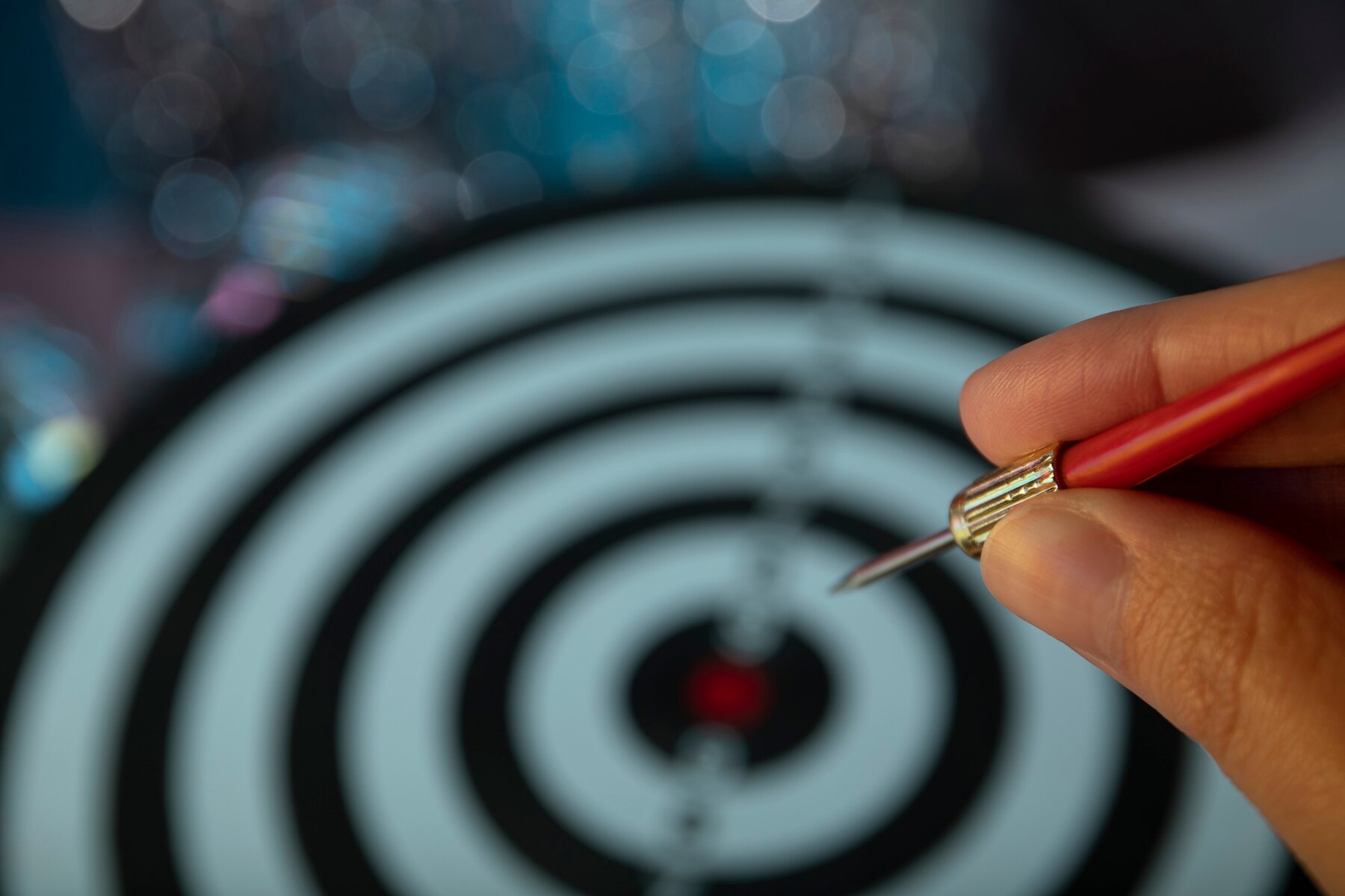 A hand holding a red dart aimed at the bullseye of a dartboard, with a blurred blue bokeh background.