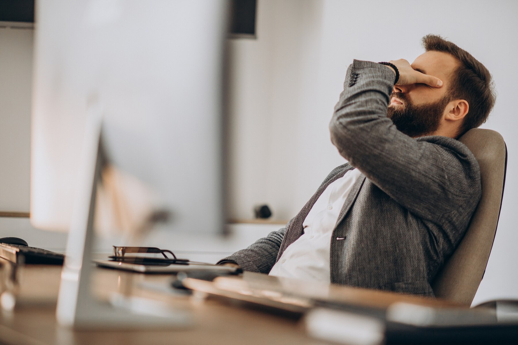 A man in a business suit sitting at a desk, appearing stressed or fatigued, with his hand covering his face.