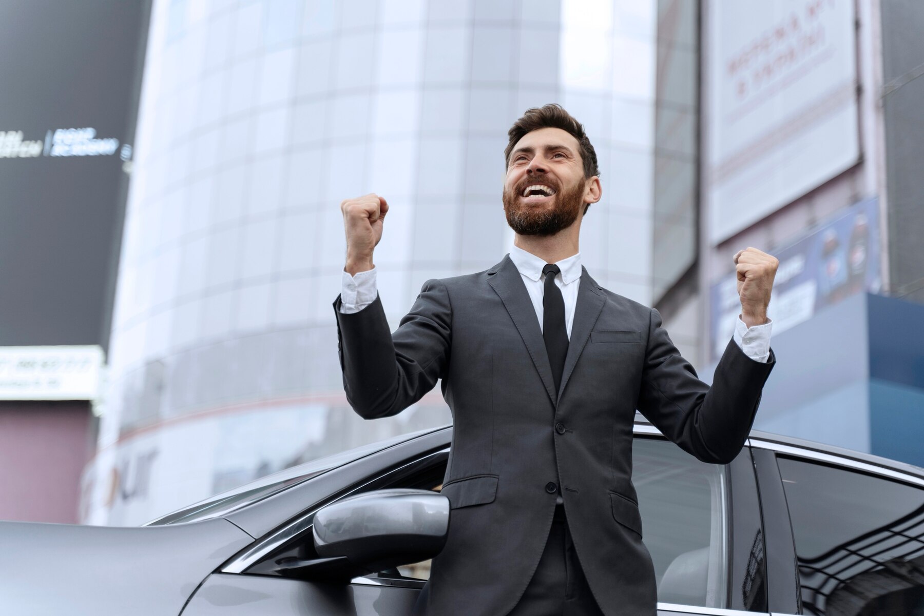 A joyful businessman in a dark suit and tie celebrating with clenched fists, standing beside a car in an urban setting with modern buildings in the background.