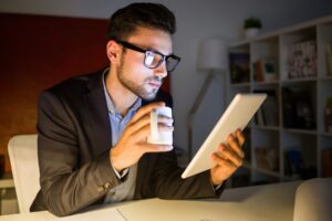 A man in a suit sits at a desk in a dimly lit room, holding a coffee cup and using a tablet, with shelves of books behind him.