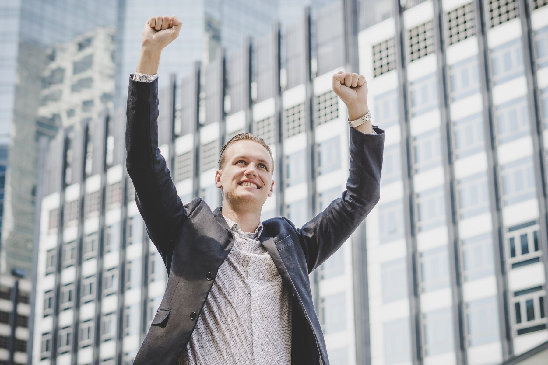 A young businessman with blond hair, dressed in a suit, celebrates a success with his arms raised in triumph, standing in front of modern glass skyscrapers.