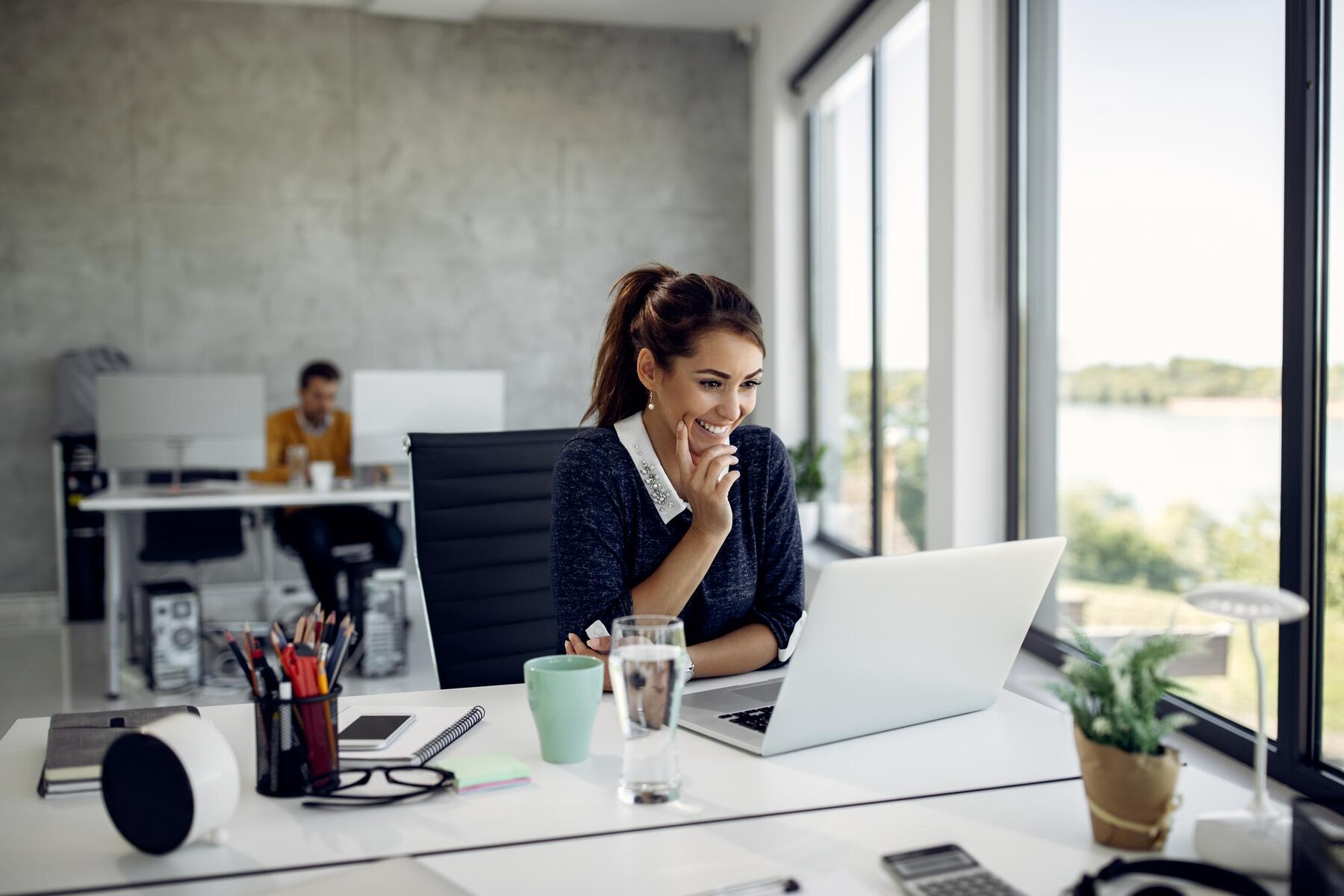 A young woman smiling while working on her laptop in a modern office space with large windows and another colleague working in the background.