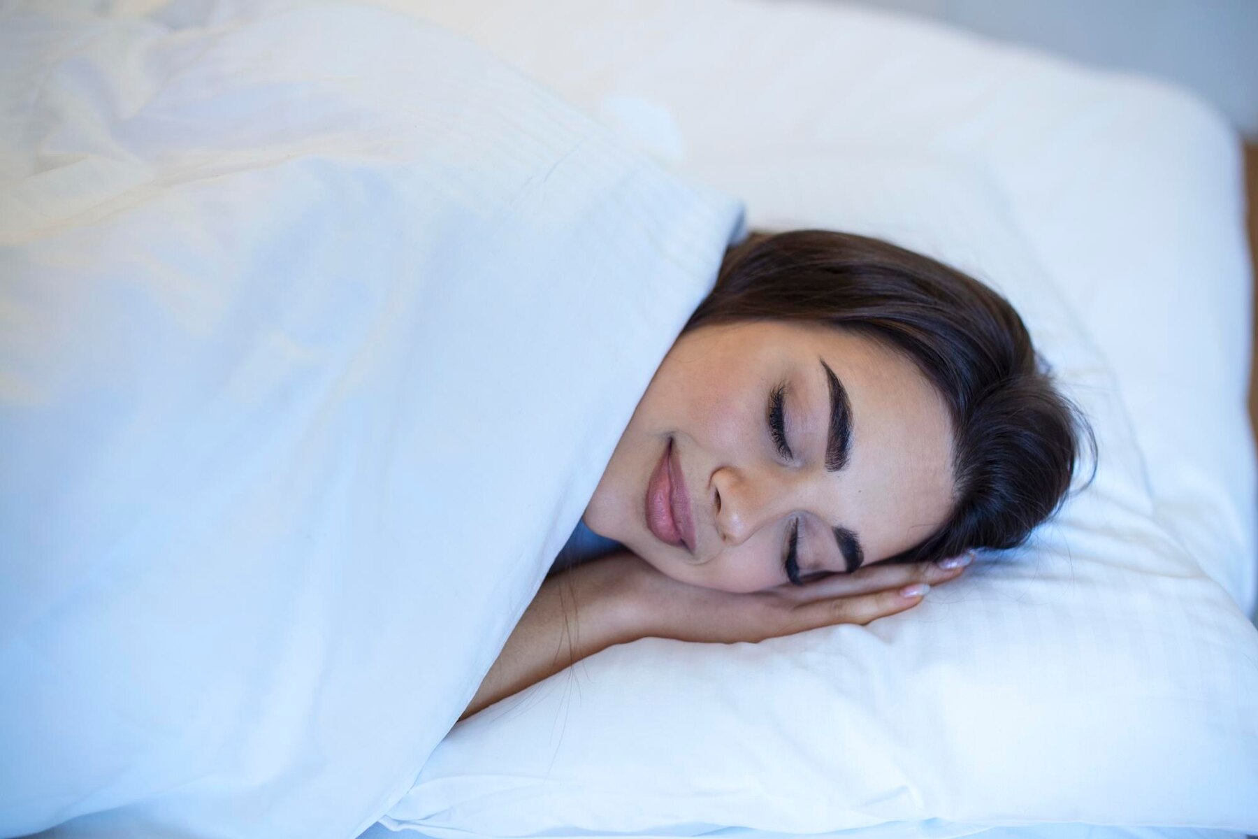 A young woman peacefully sleeping with her head resting on her hand, covered by a white duvet in a softly lit bedroom.