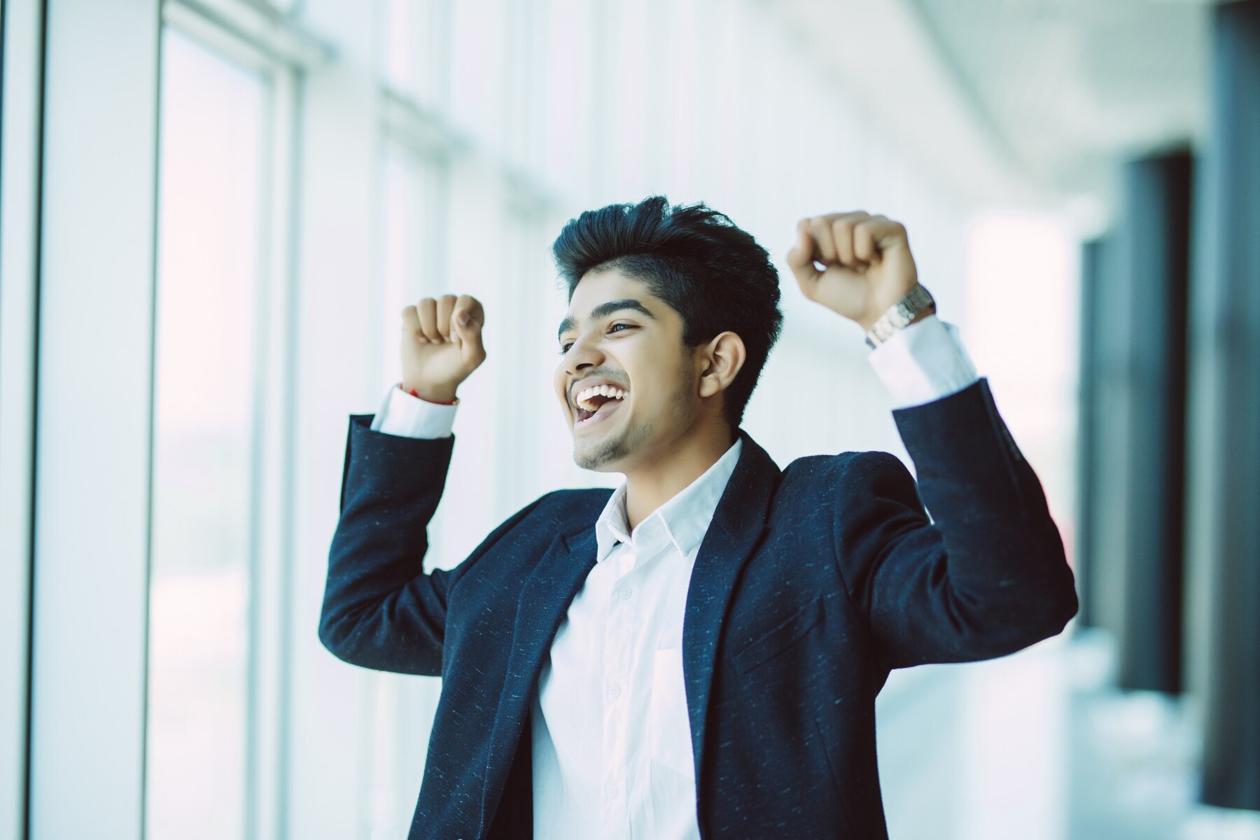 A young man in a dark suit and white shirt, joyfully raising his arms in a victory gesture, laughs and looks upwards in a bright, window-lined corridor.