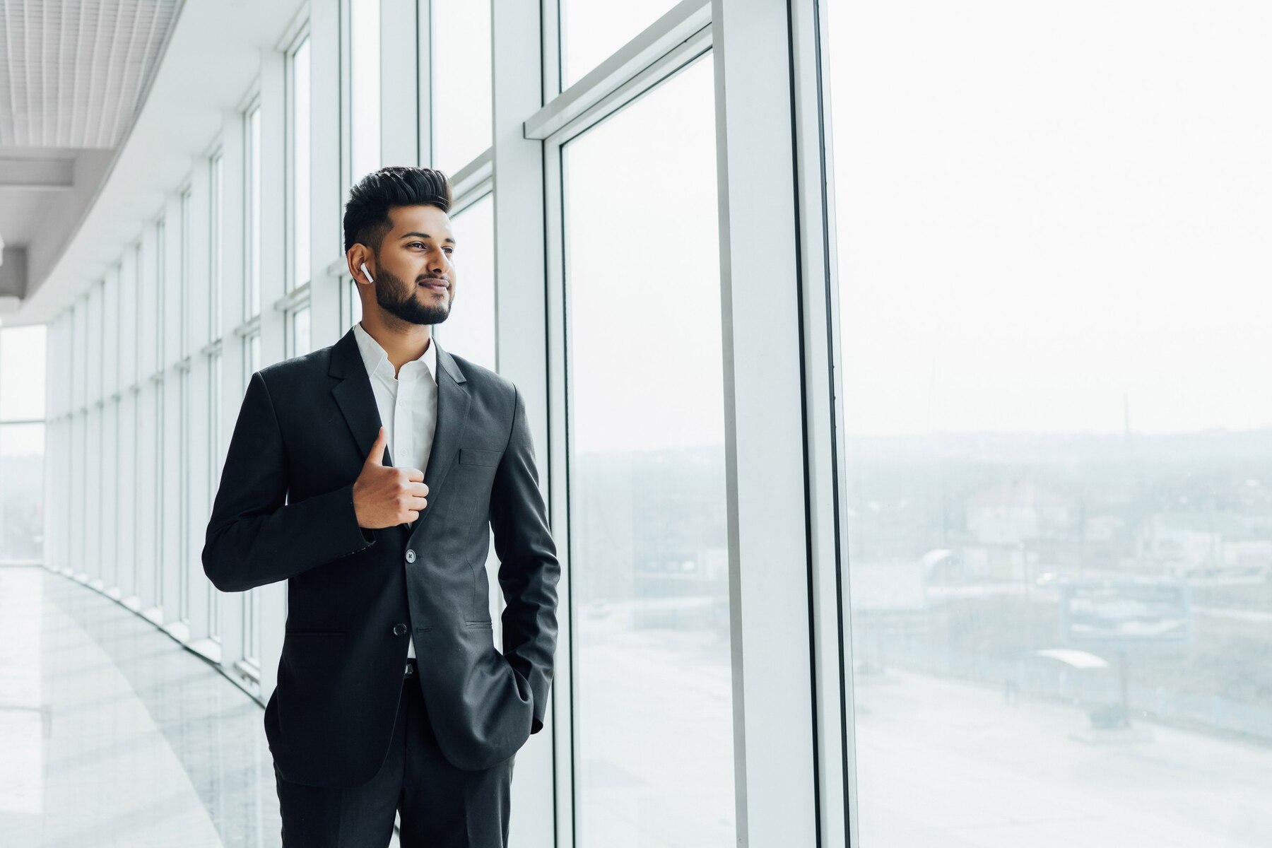 A confident young businessman in a black suit and white shirt standing by large windows in a bright modern office, using a wireless earpiece.