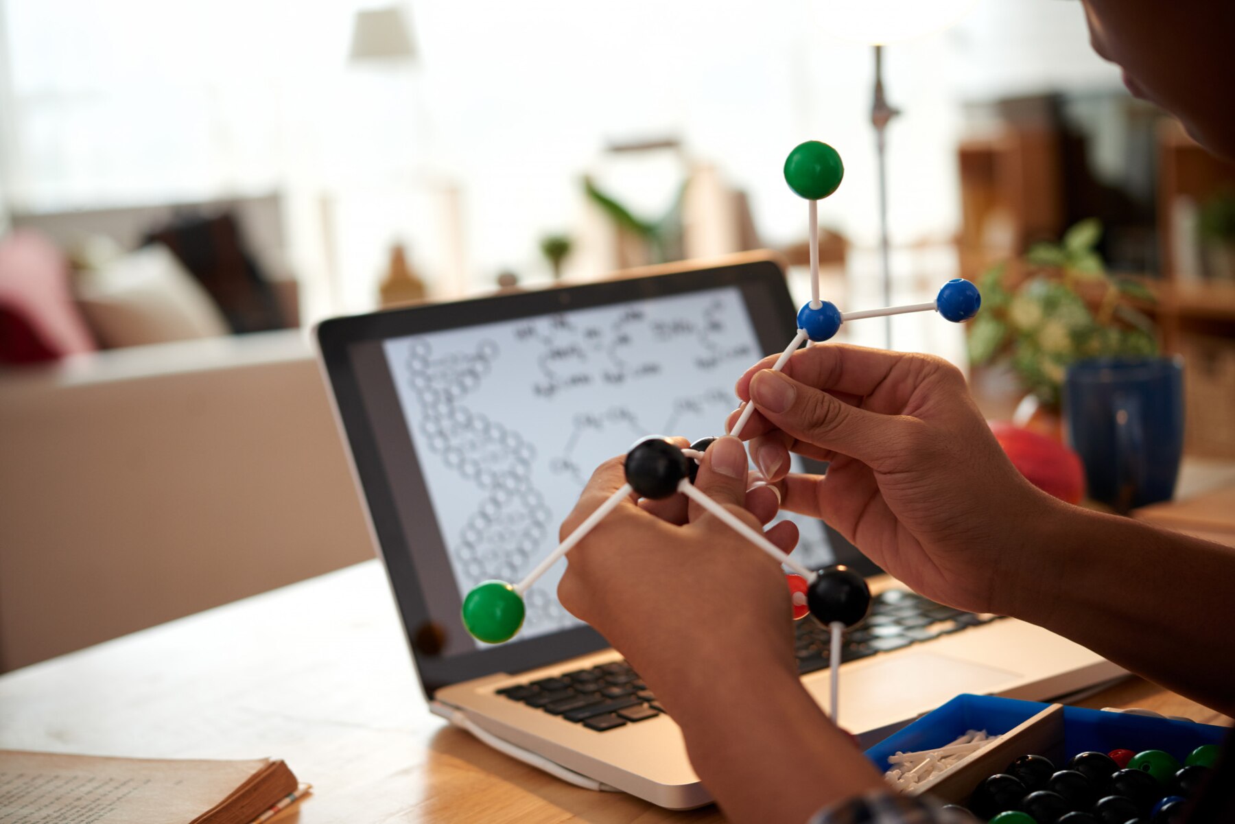 Close-up of hands assembling a molecular model with a laptop in the background displaying chemical structures, in a well-lit home office setting.