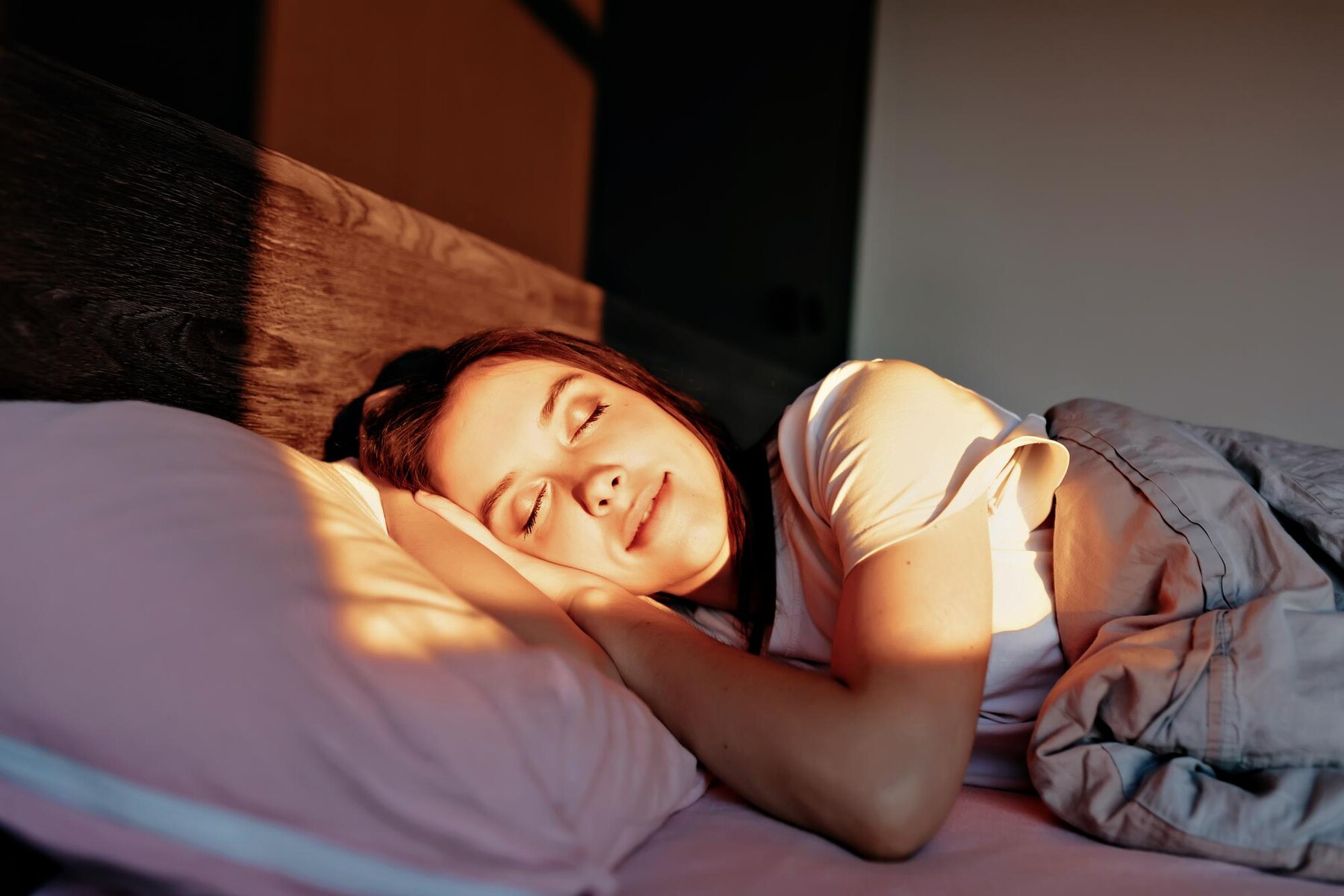 A young woman sleeps peacefully, basked in warm sunlight filtering through a window, resting her head on her arm on a bed with soft pink and grey bedding.