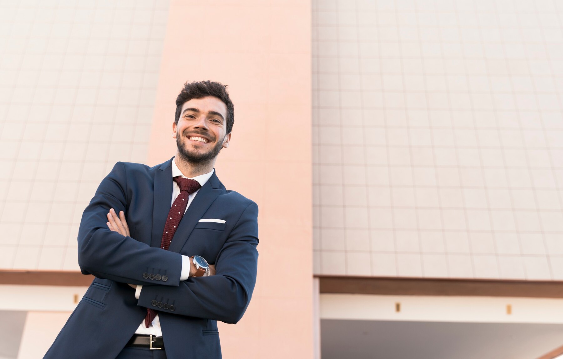 A confident young businessman in a navy suit with arms crossed, smiling outdoors with a light pink building background.