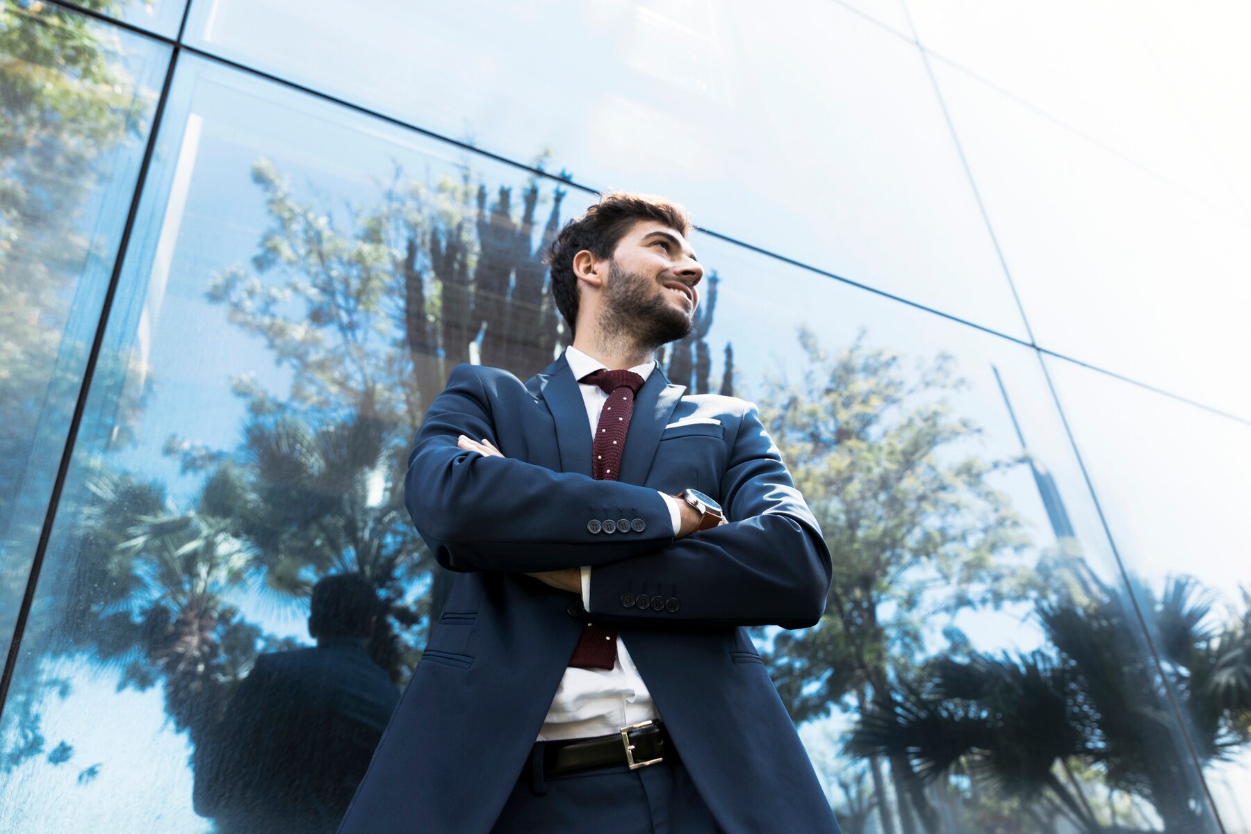 Confident young businessman in a blue suit with crossed arms looking upwards, standing near a reflective glass building under a clear blue sky.