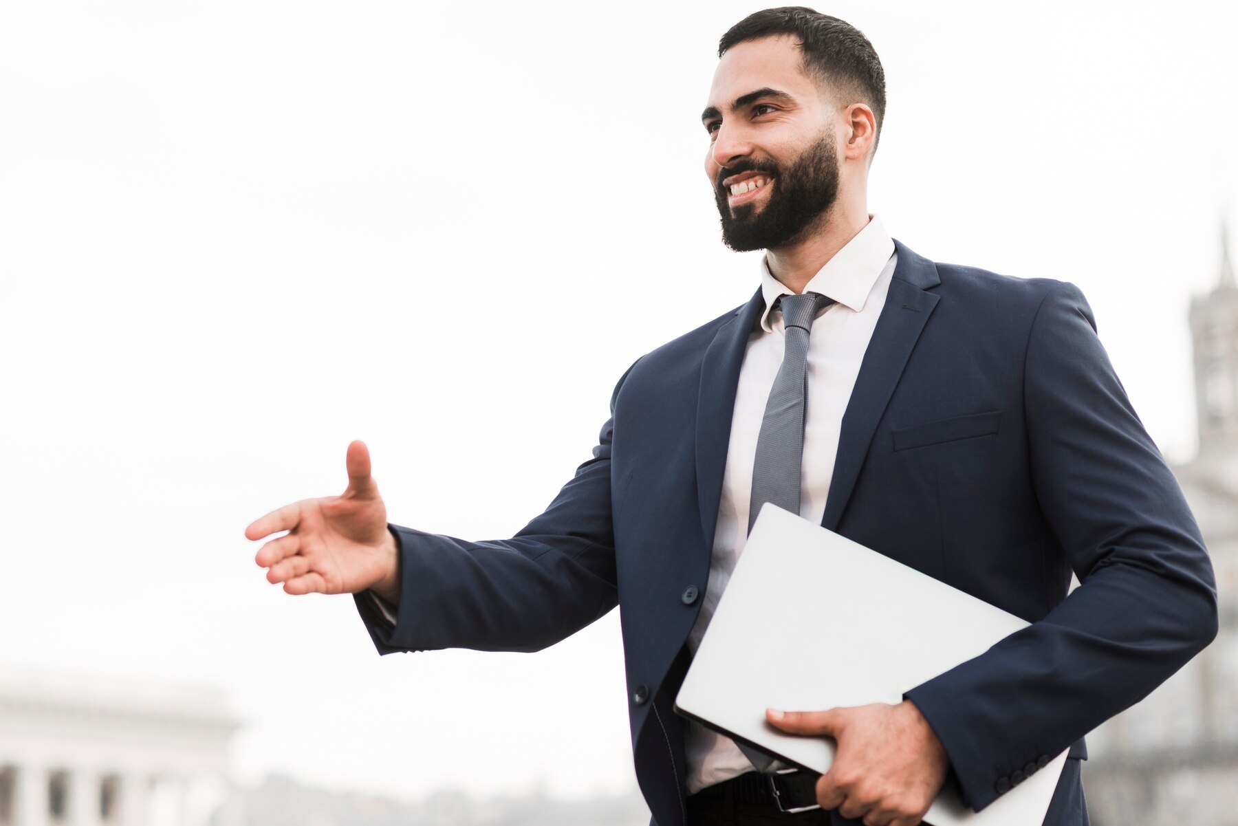 A professional man in a suit with a beard, smiling and extending his hand in greeting, holding a laptop under his arm, in an urban environment.
