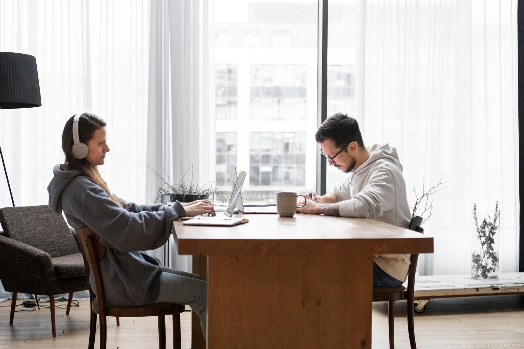 Two individuals work collaboratively at a wooden table, laptops open, in a bright room with large windows and indoor plants.
