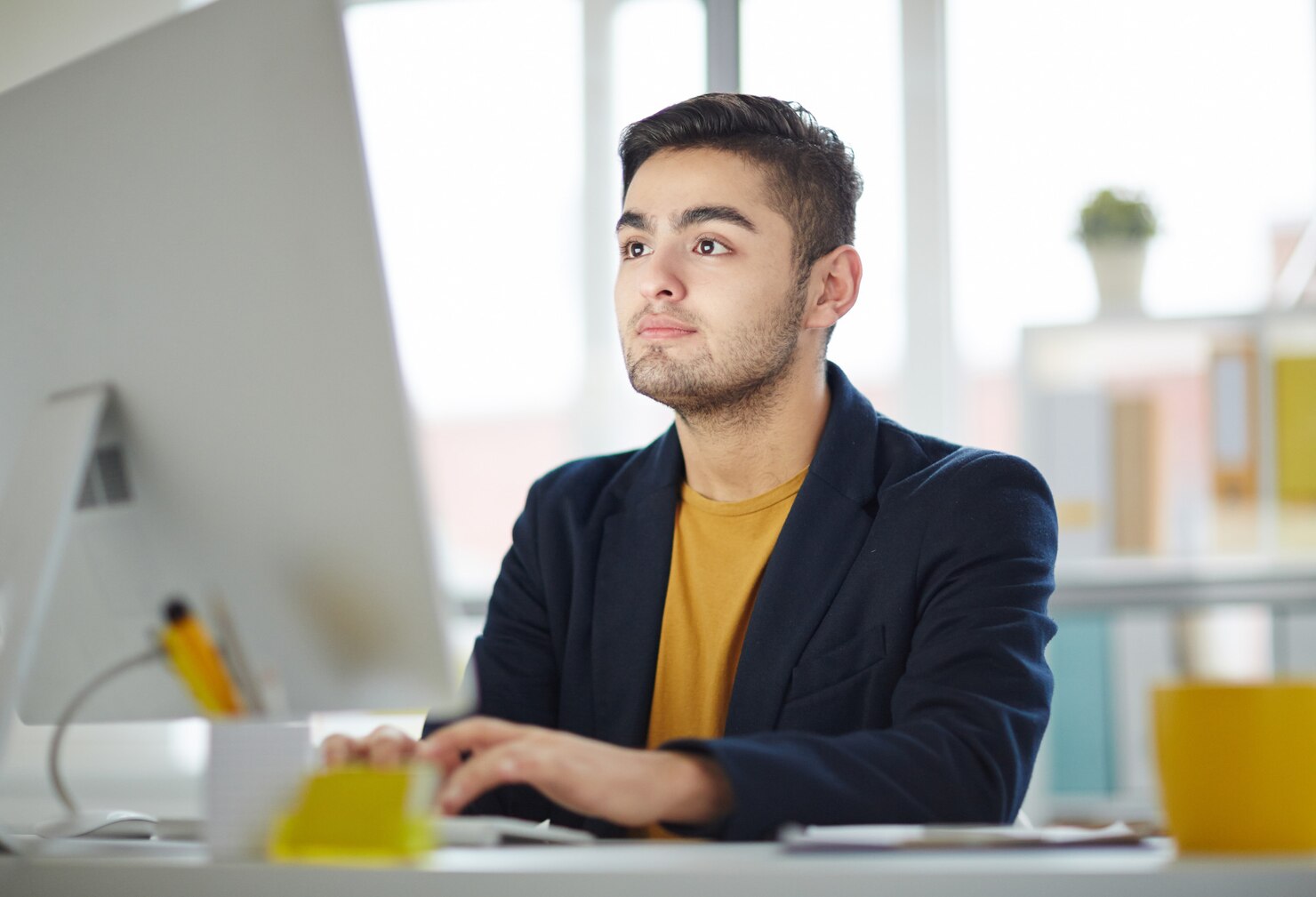 A person in a navy blazer and yellow shirt is working at a desk with a computer, surrounded by colorful office supplies and soft lighting.