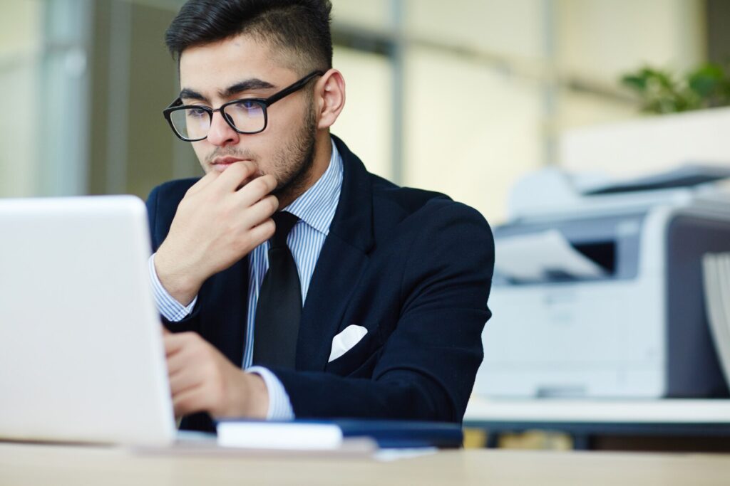 A person in a suit sits at a desk, thoughtfully engaged with a laptop, surrounded by an office environment and printer.
