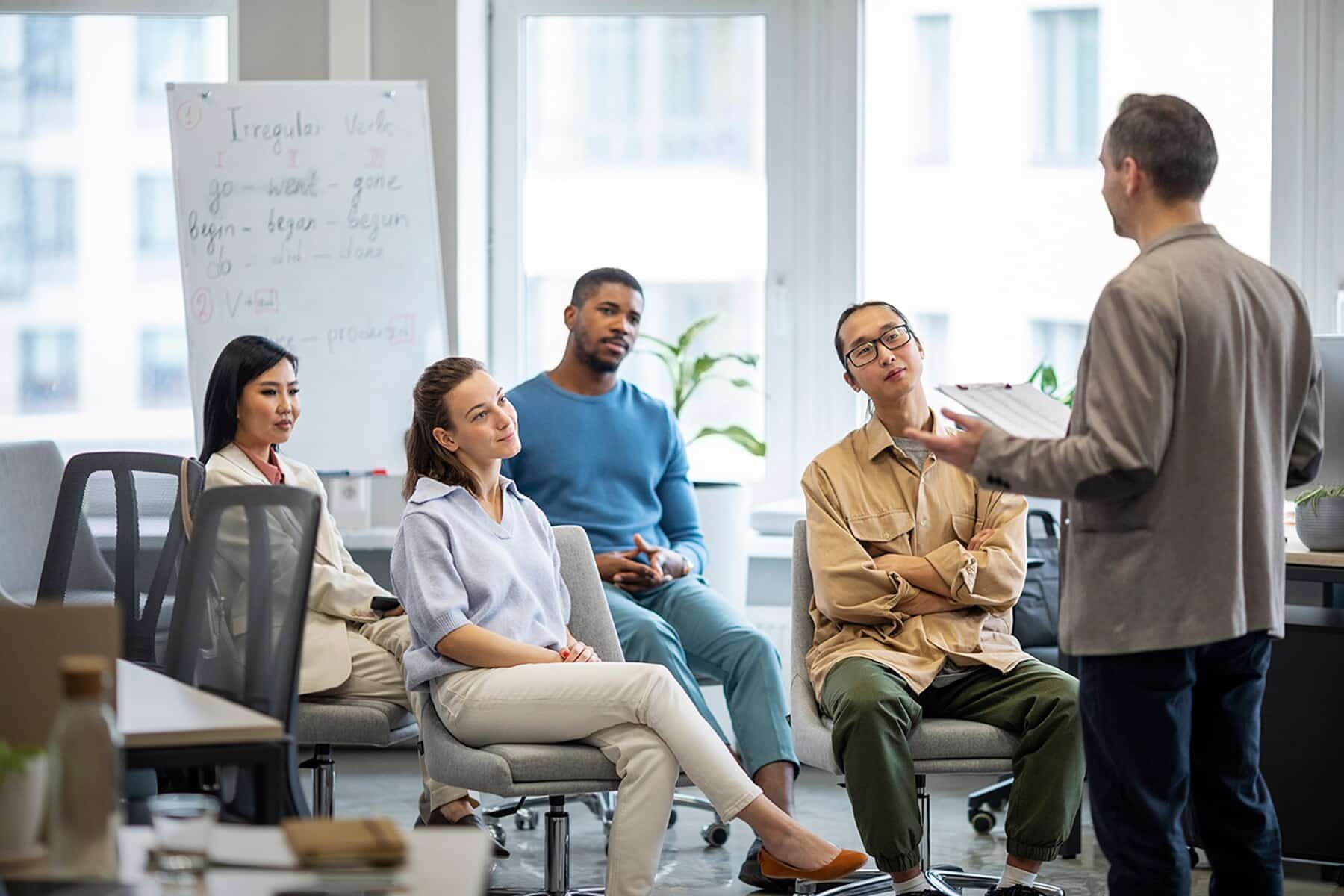A group of four people attentively listens to a speaker in a bright office space, with a presentation on the whiteboard behind them.