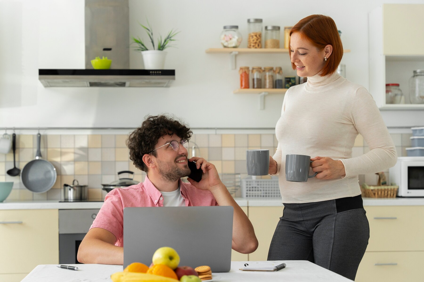 A man with curly hair is talking on the phone and smiling at a red-haired woman who is holding two grey mugs while they stand in a modern kitchen.